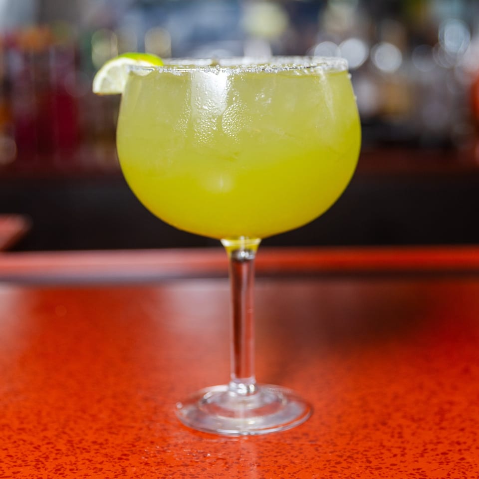 Refreshing green margarita with a salted rim and lime wedge on a red bar top.