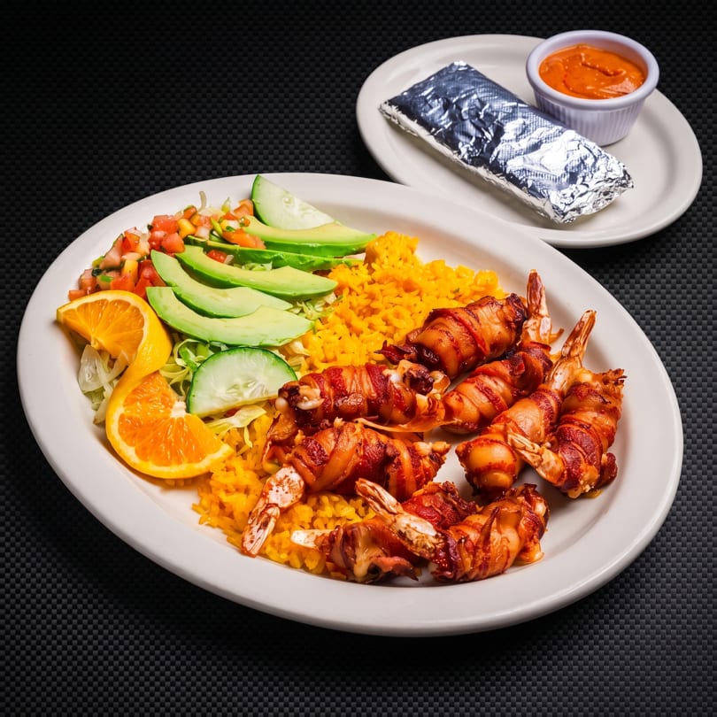 Plate of bacon-wrapped shrimp, yellow rice, salad, and orange, with a side of sauce and foil-wrapped item.