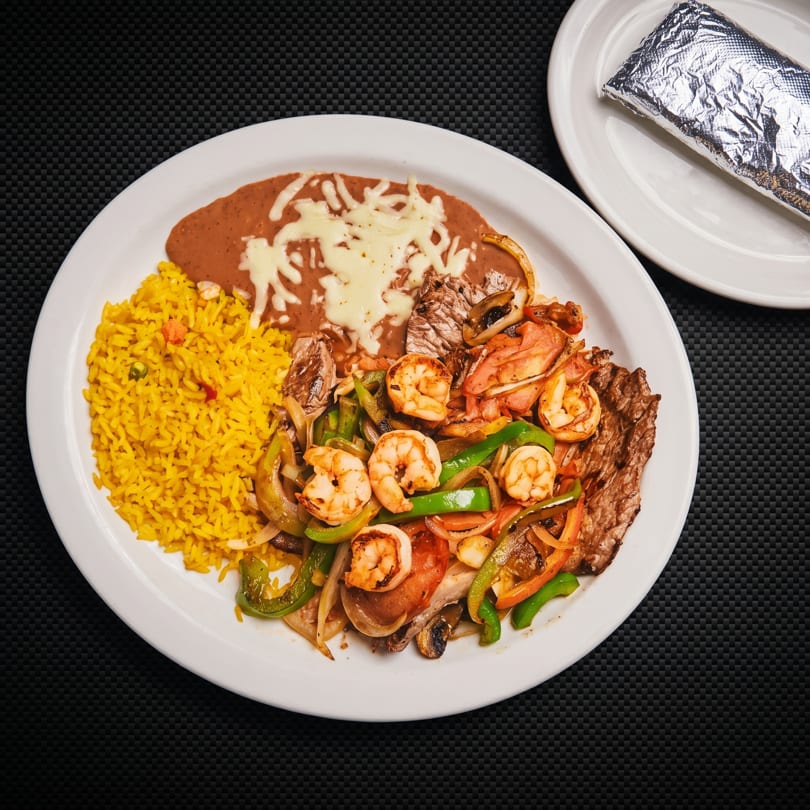 Fajita plate with yellow rice, refried beans, steak, shrimp, vegetables, and foil-wrapped tortillas.