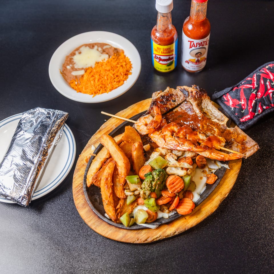 Sizzling steak and shrimp fajita platter with fries, vegetables, rice, beans, and hot sauce.