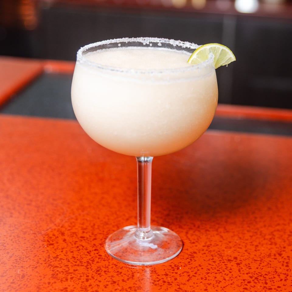 Frozen margarita in a stemmed glass with a salt rim and lime wedge on a red bar.