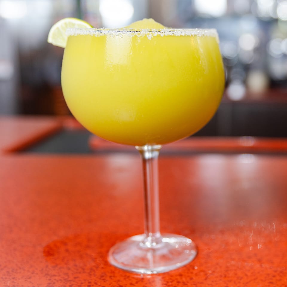Bright yellow frozen margarita with a salt rim and lime wedge on a vibrant red bar.