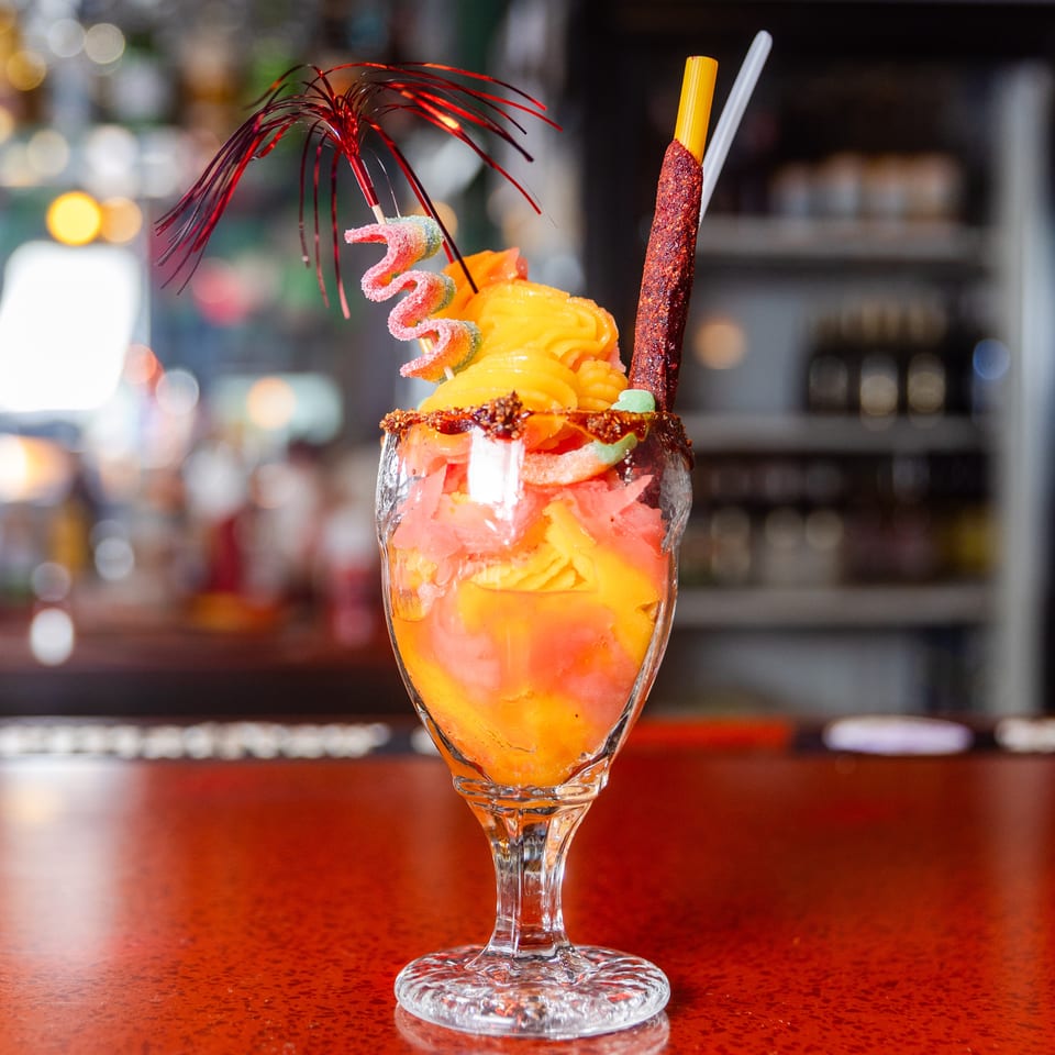 Colorful mango dessert drink with gummies, a red umbrella, and a chili-dusted straw on a bar.