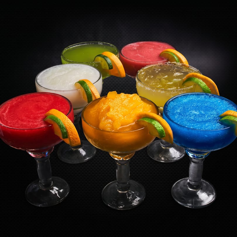 Seven colorful frozen margaritas garnished with orange and lime slices on a dark background.