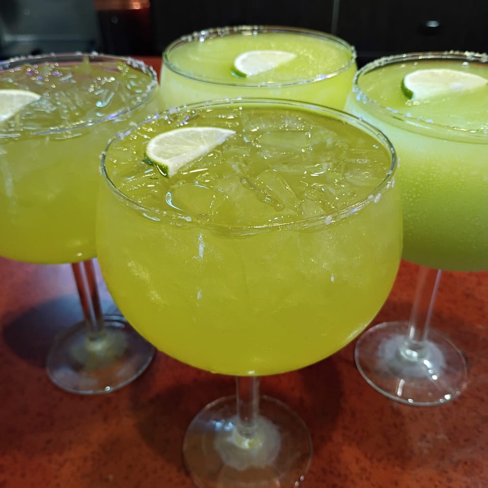 Four glasses of vibrant green margaritas with salt rims and lime wedges on a reddish-brown table.